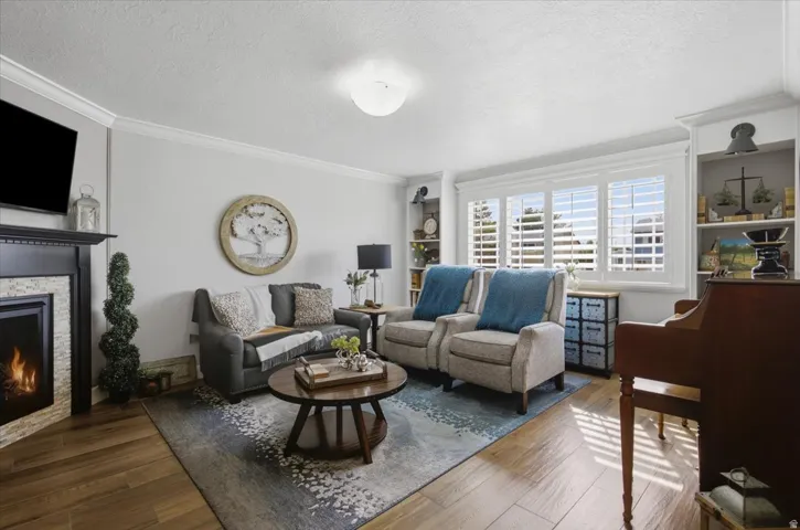 Wood-finish flooring, built-in shelving, white plantation shutters, corner fireplace with stone surround, and crown molding