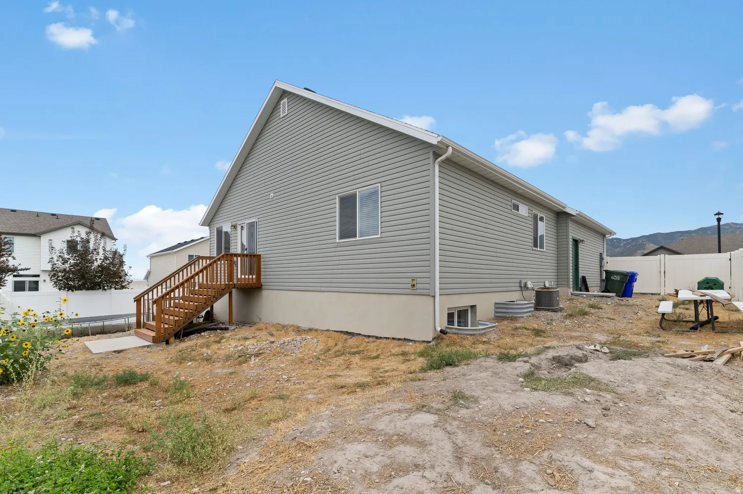 Rear exterior of the residence featuring light gray siding, a wood-finish deck with stairs, a white vinyl fence, and foundation window wells