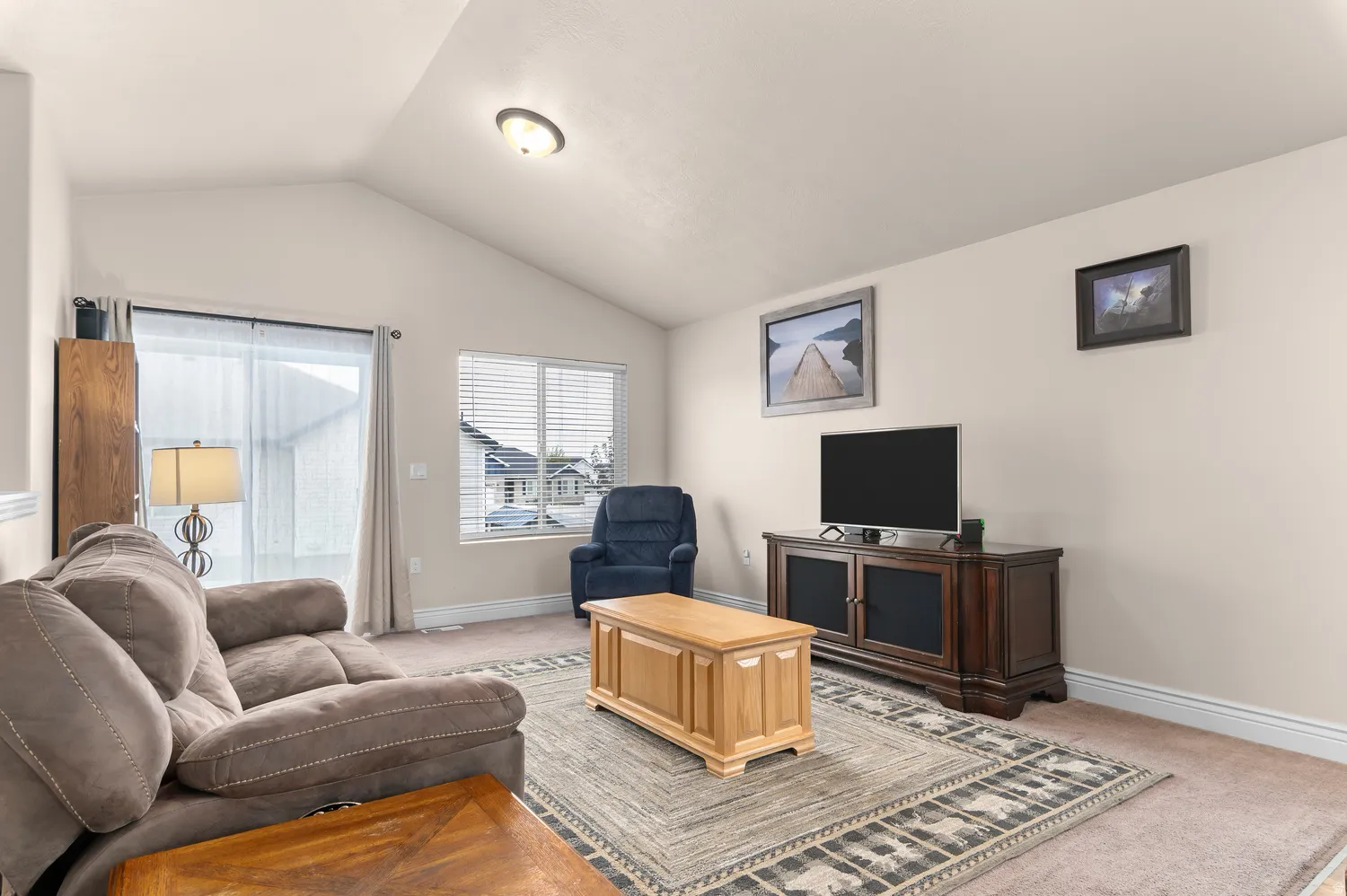 Vaulted ceiling living space with carpet flooring, a large window with blinds, and a sliding glass door