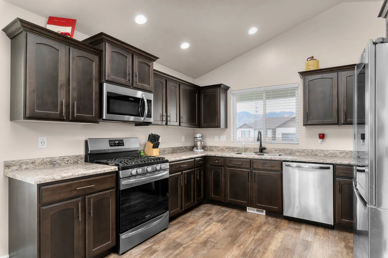 Kitchen with wood-finish flooring, dark wood cabinetry, stainless steel appliances, speckled countertops, and a vaulted ceiling