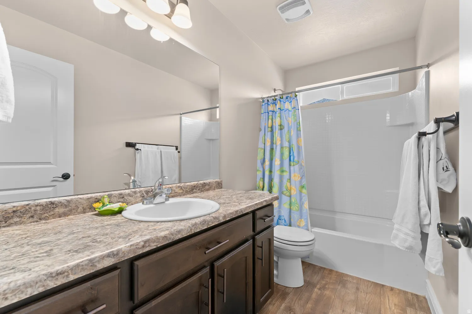 Full vanity mirror, integrated sink, wood-finish flooring, dark wood cabinetry, and a full bathtub with shower