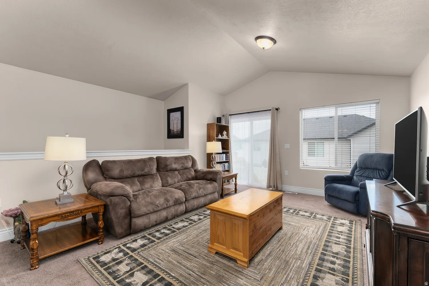 Vaulted ceilings, neutral wall tones, carpeted flooring, and a sliding glass door with blinds