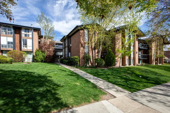 Brick exterior buildings with multiple balconies, surrounded by mature trees and manicured lawns, featuring concrete walkways