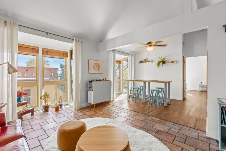 Vaulted white ceilings, expansive window wall with wooden blinds, ceramic tile flooring, wood-finish flooring, and a ceiling fan with light fixture