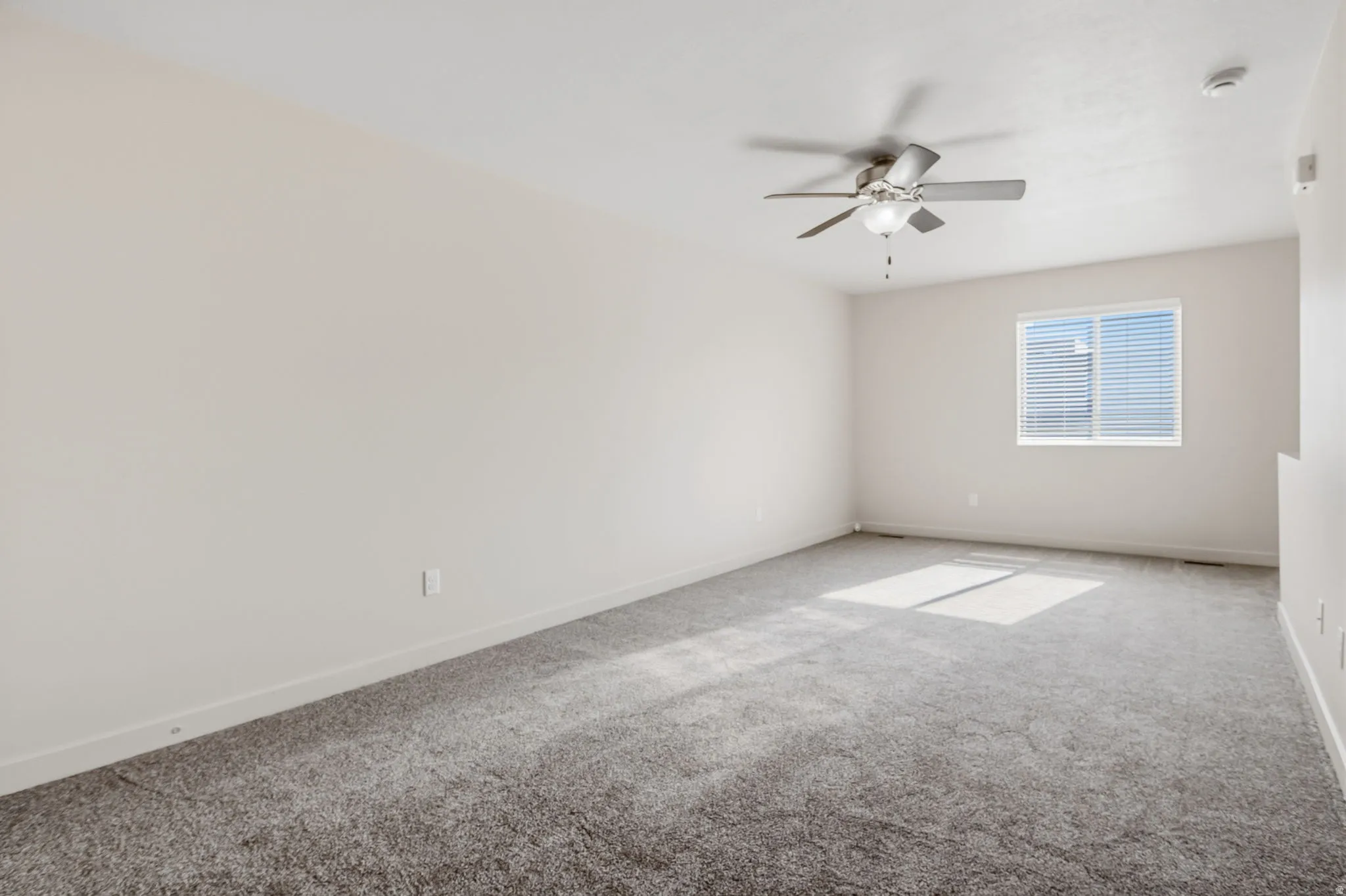 Plush carpet flooring, neutral wall tones, window with horizontal blinds, and a ceiling fan with integrated lighting