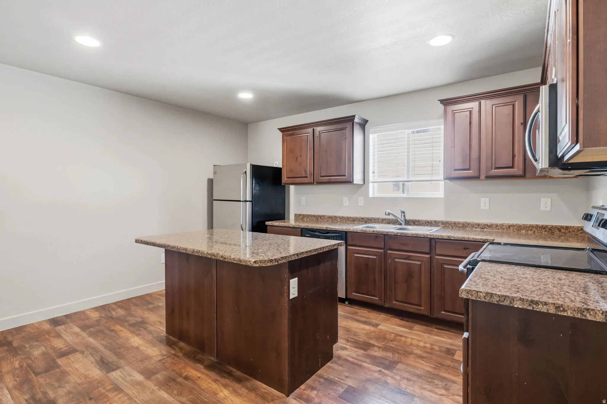 Kitchen featuring wood-finish flooring, dark wood cabinetry, granite-style countertops, an island with extended counter space, and stainless steel appliances