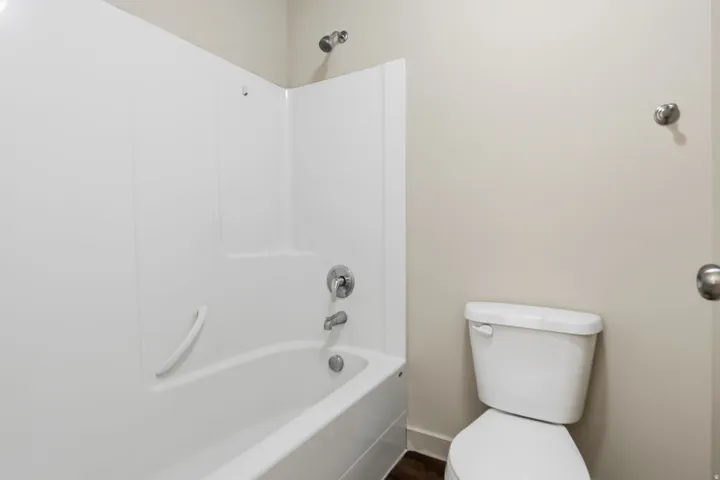 White tub with integrated shower surround, chrome plumbing fixtures, white toilet, and neutral wall paint