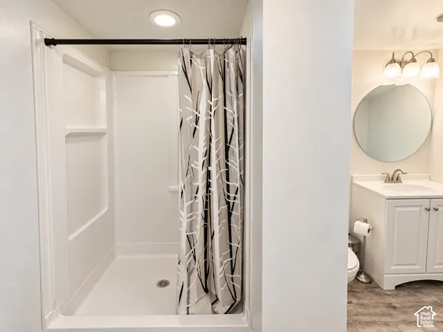 Bathroom featuring a walk-in shower with integrated surround, wood-finish flooring, white cabinetry with countertop-integrated sink, and a circular wall-mounted mirror with a three-light vanity fixture