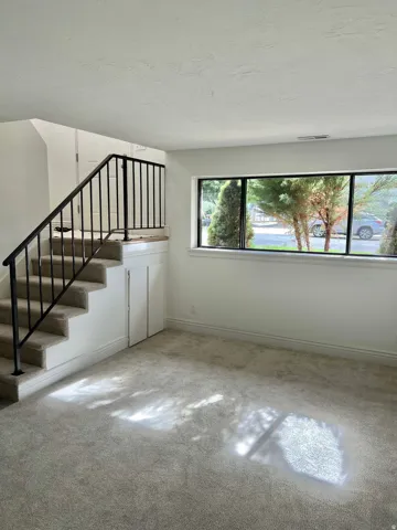 Carpeted flooring, white walls, black metal staircase railing, under-stair storage, and a large multi-pane window
