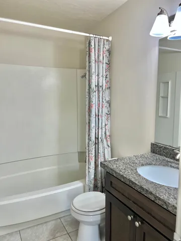 Integrated bathtub and shower combination with white surround, tile flooring, dark wood-finish vanity with stone countertop, and single basin sink