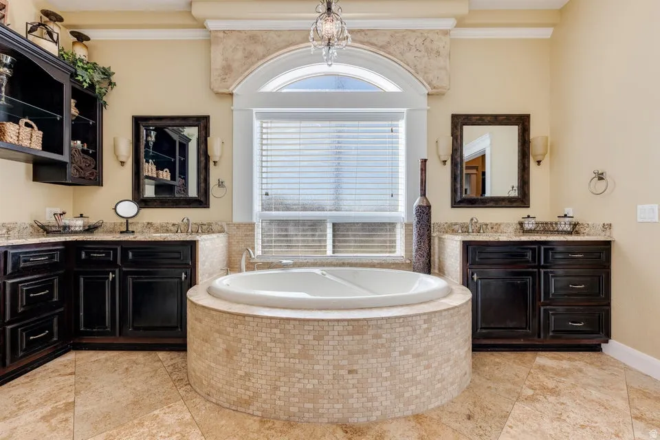 Built-in soaking tub with mosaic tile surround, dual vanity areas with dark wood cabinetry and stone countertops, arched window with privacy blinds, and tile flooring