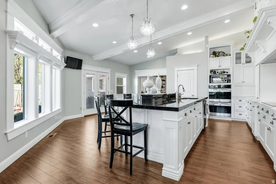 Kitchen featuring wood-finish flooring, a central island with a dark countertop, white cabinetry, integrated double ovens, and vaulted ceilings with exposed beams