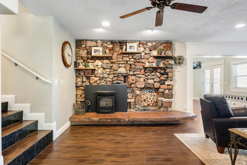 Stone fireplace with a wood-burning insert, wood-finish flooring, staircase with black-finish risers, ceiling fan, and recessed lighting
