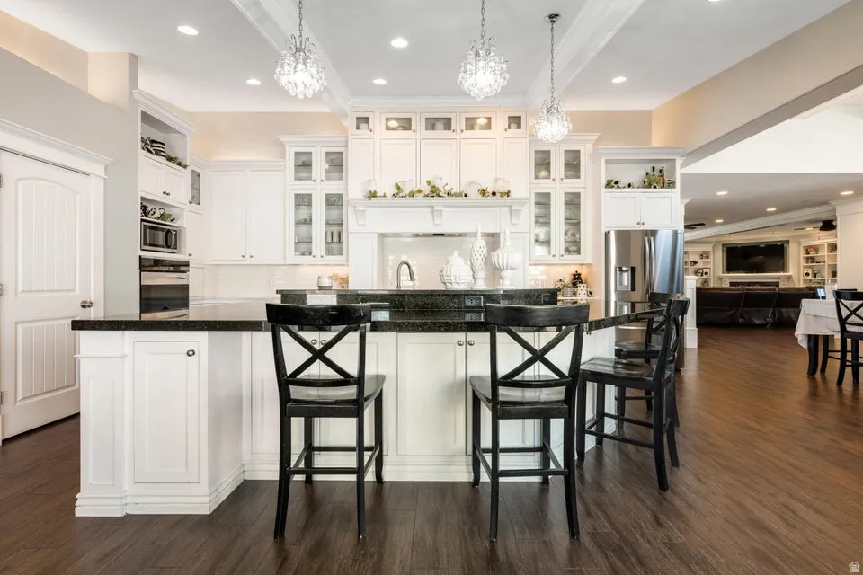 White cabinetry with glass-front accents, dark countertops, wood-finish flooring, recessed lighting, and crystal chandeliers