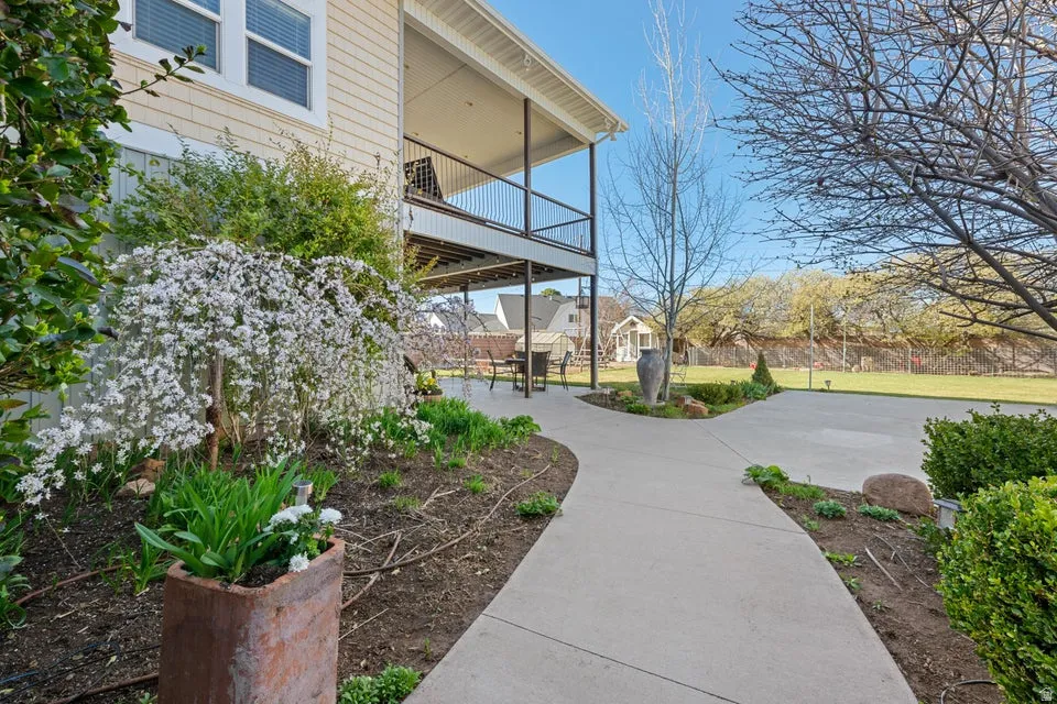 Exterior featuring light-colored siding, a covered upper-level balcony, a lower-level patio, a concrete pathway, and landscaped garden beds