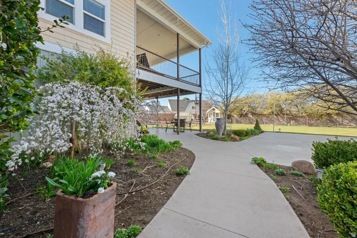 Exterior featuring light-colored siding, a covered upper-level balcony, a lower-level patio, a concrete pathway, and landscaped garden beds