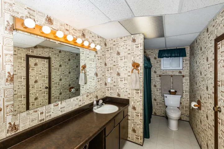 Spa-inspired bathroom featuring custom tile walls with decorative mosaic detailing, a large framed mirror with integrated vanity lighting, dual sink setup with dark wood cabinetry, and a separate water closet with window for natural light