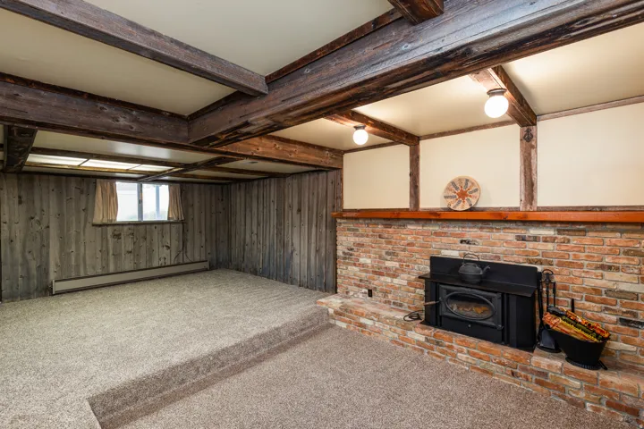 Spacious basement recreation area featuring exposed wooden beam ceiling, brick fireplace with black insert stove, carpeted flooring with raised platform section, and recessed lighting throughout