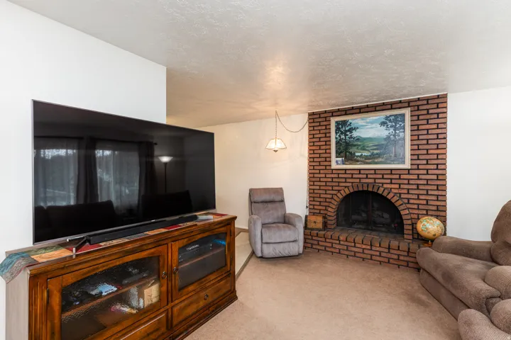 Comfortable family room featuring a brick fireplace with hearth, wood-finish media console with glass-front storage, pendant lighting, plush seating areas, and neutral carpeting throughout
