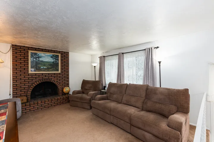Spacious living room featuring a brick fireplace with decorative mantel, large picture window with natural light, neutral carpeting, and ceiling-mounted lighting fixtures