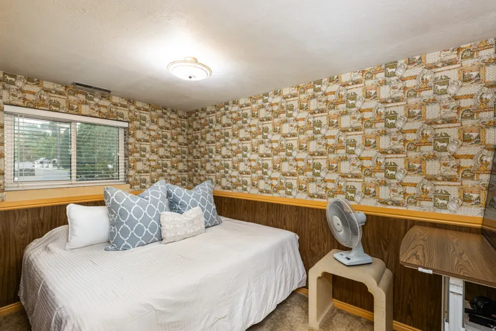 Cozy bedroom featuring warm wood-finish wainscoting, patterned wallcovering, and a picture window with natural light