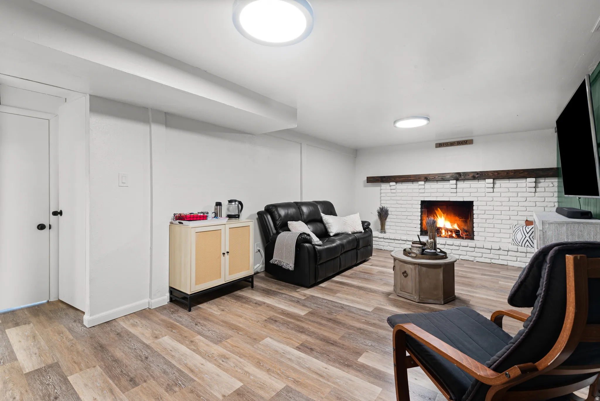 Whitewashed brick fireplace with a wood mantle, wood-finish flooring, and recessed ceiling lighting