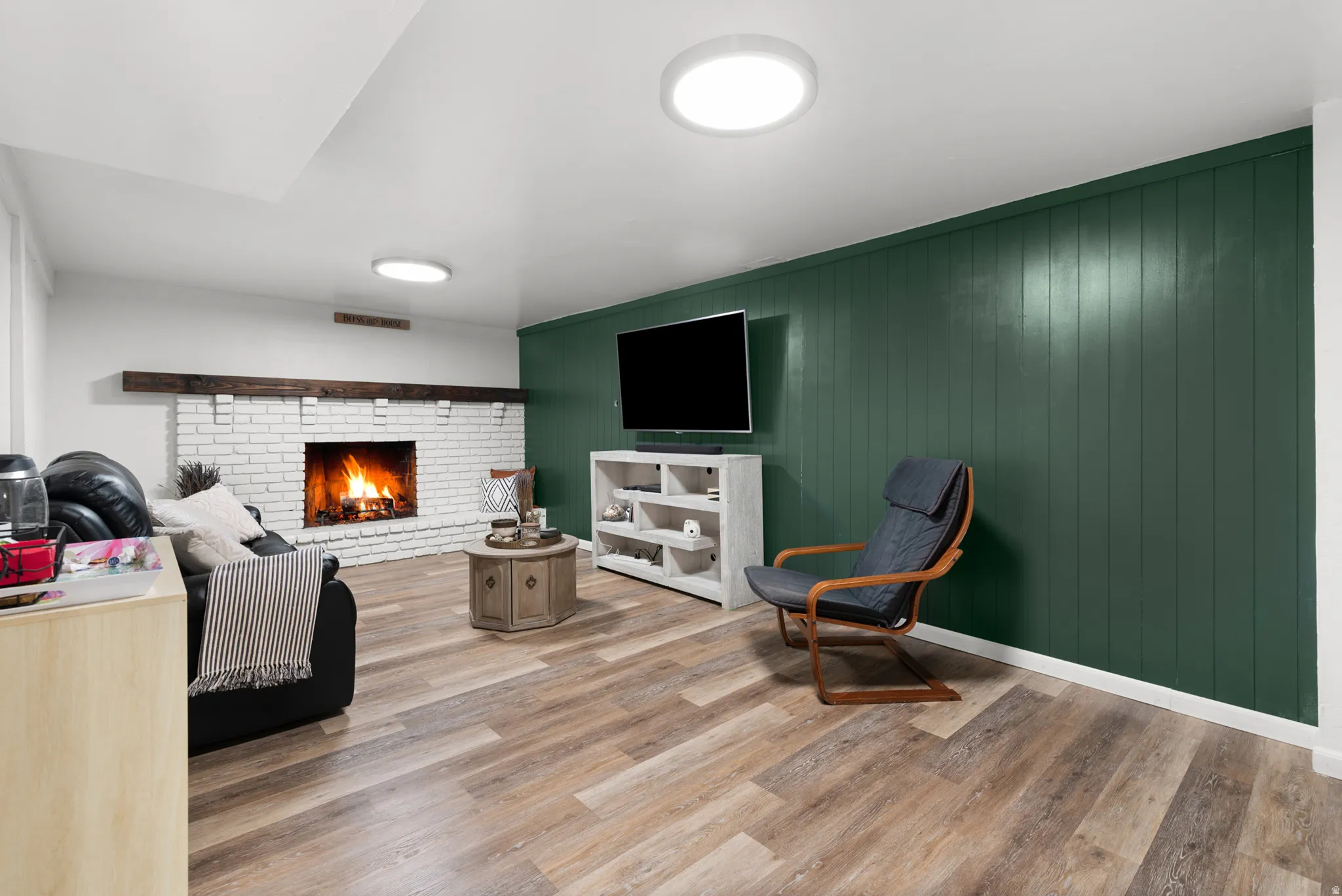 Finished basement with Warm wood-finish flooring complements a white-painted brick fireplace with a substantial wood mantel