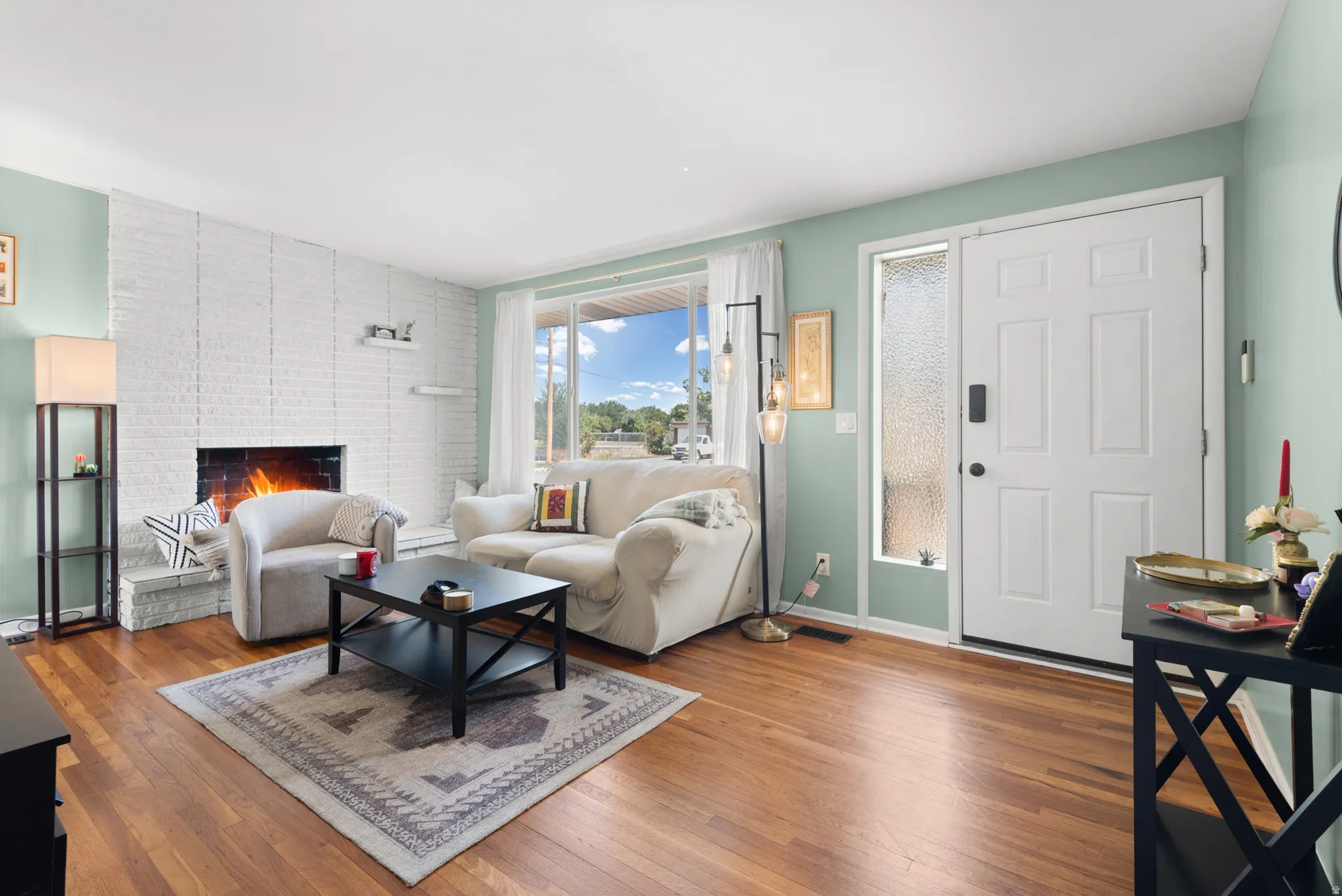 Hardwood flooring, white brick fireplace, expansive window, white paneled entry door, and textured glass sidelight