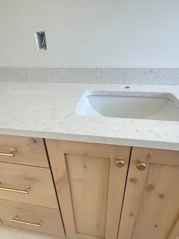 Light wood-finish vanity cabinetry with gold-tone hardware, an integrated undermount sink, and a speckled light-toned countertop with matching backsplash