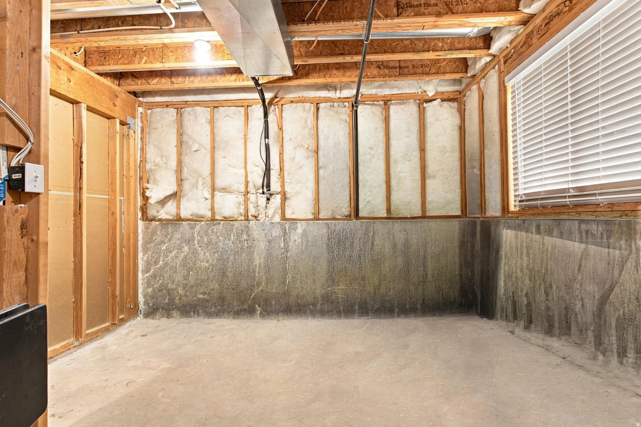 Unfinished lower level featuring exposed wood framing, concrete flooring, and a window with horizontal blinds