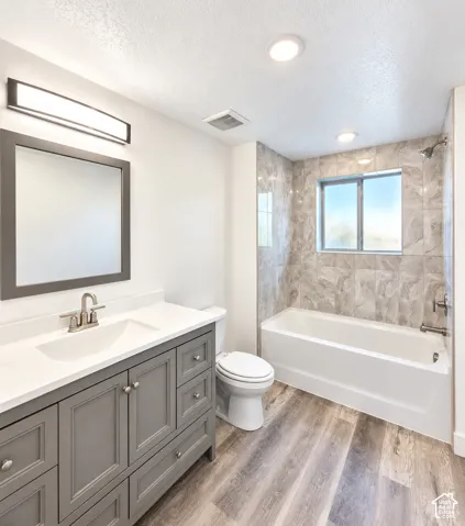 Gray wood-finish flooring, built-in vanity with white countertop, recessed lighting, chrome fixtures, and tiled tub/shower combination