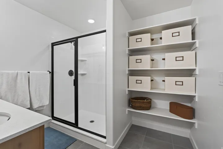 Main floor owners bathroom with shower with dark-framed glass enclosure, built-in shelving, vanity with light-toned countertop, and recessed lighting.