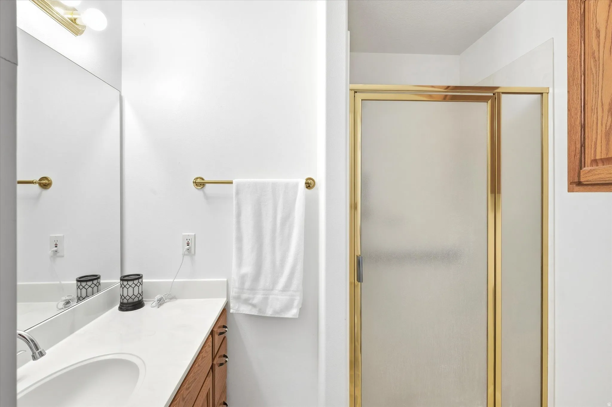 Bathroom featuring a wood-finish vanity with a light-toned countertop, an integrated sink, a large wall mirror, and a shower with a gold-toned framed frosted glass enclosure
