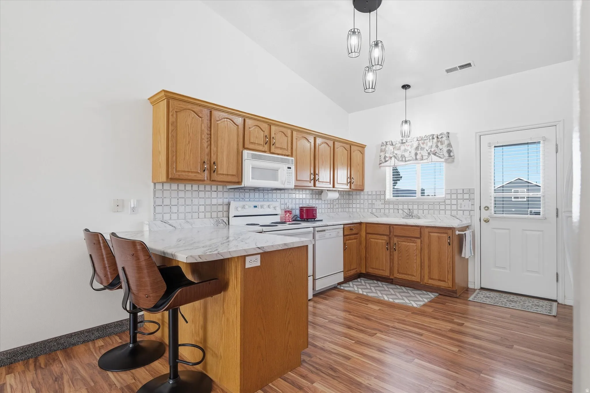 Vaulted ceiling kitchen featuring wood-finish cabinetry, a breakfast bar with stone-finish countertops, white appliances, and a tiled backsplash