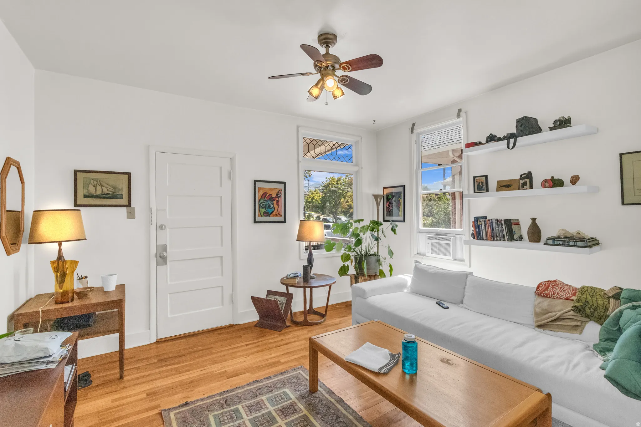 Hardwood flooring, crisp white walls, multiple windows, a ceiling fan with integrated lighting, and built-in floating shelves