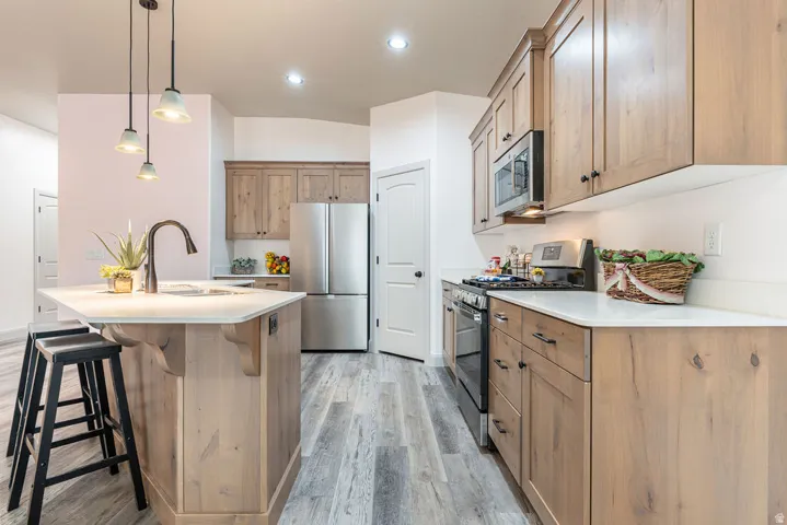Kitchen featuring wood-finish cabinetry, stainless steel appliances, a center island with sink, pendant lighting, and light-toned flooring