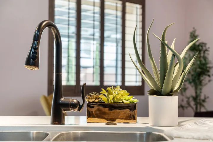 Matte black pull-down faucet over a double basin sink, set within a light-toned countertop, with plantation shutters in the background