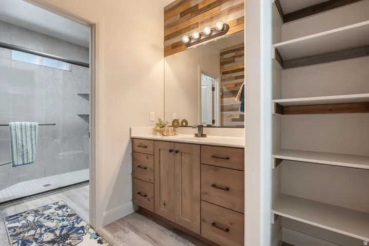 Wood-finish vanity with a light-toned countertop, a multi-paneled wood accent wall, built-in shelving, and a glass-enclosed shower with tile surround