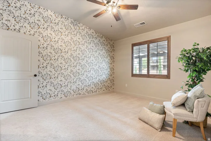Textured wallpaper accent wall, wood-frame window with plantation shutters, ceiling fan with integrated lighting, paneled interior door, and neutral-tone carpeting