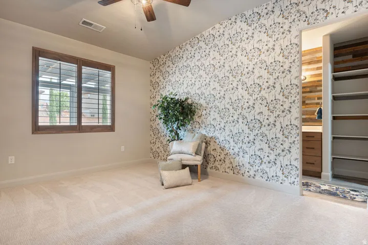 Wall-to-wall carpeting, wood-frame window with plantation shutters, ceiling fan, accent wall with floral wallpaper, and built-in closet shelving