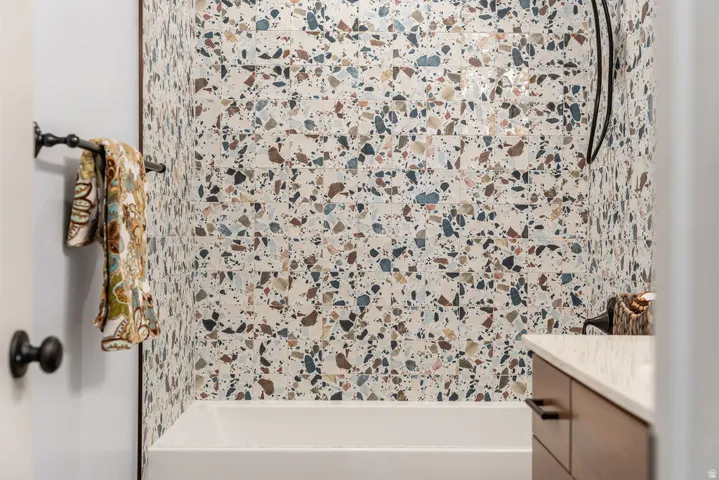 Terrazzo-style tile shower surround with a white tub, dark metal shower fixtures, and a wood-finish vanity with a light-colored countertop