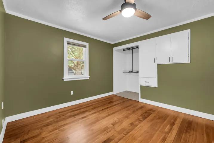Spacious room featuring warm wood-finish flooring, sage green walls with crisp white trim, a ceiling fan with integrated lighting, a large window with white frame, and built-in white cabinetry with storage and hanging rod