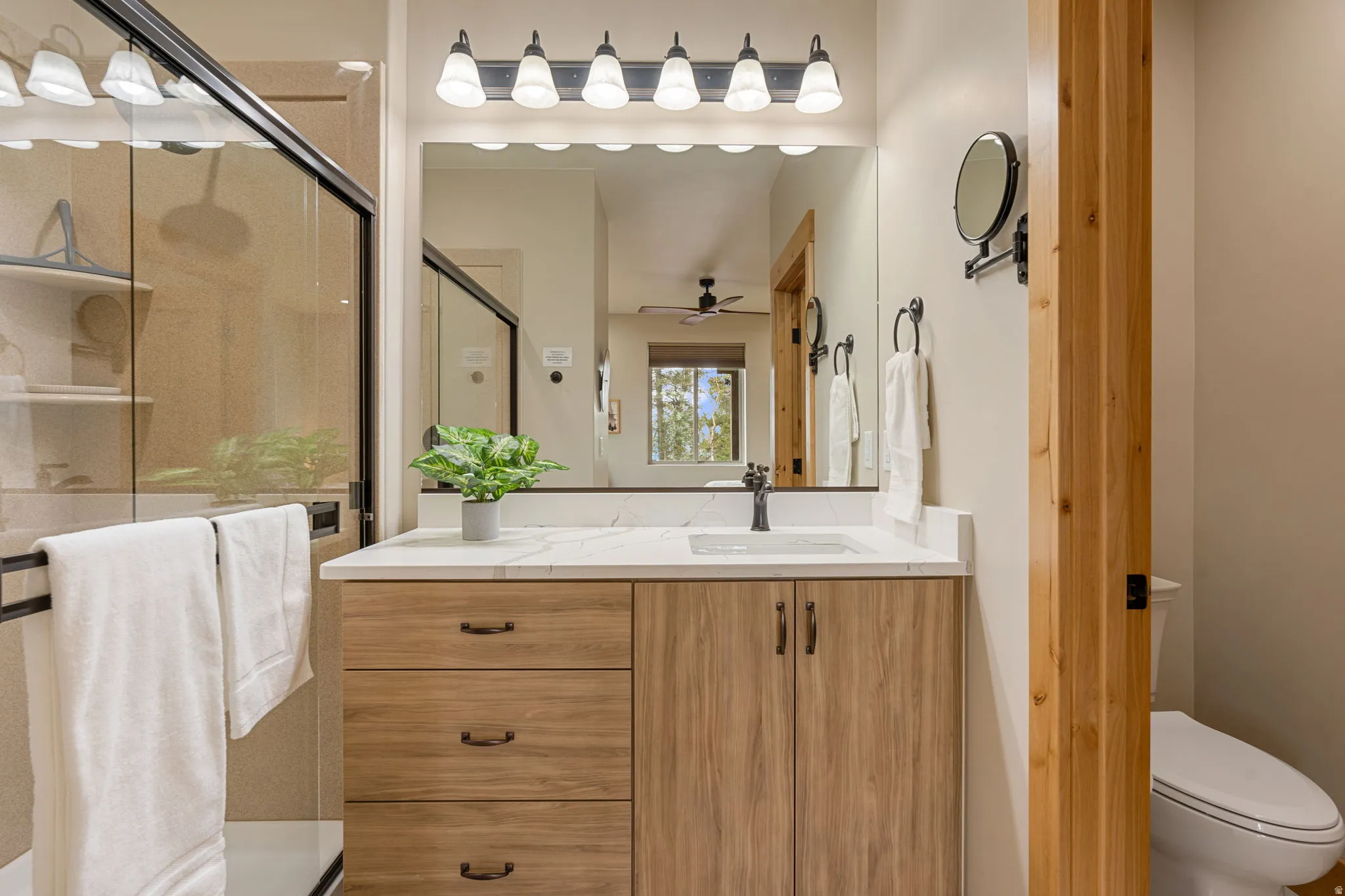Wood-finish vanity with a white countertop and integrated sink, framed mirror with a six-light vanity fixture, glass-enclosed shower, and a built-in toilet
