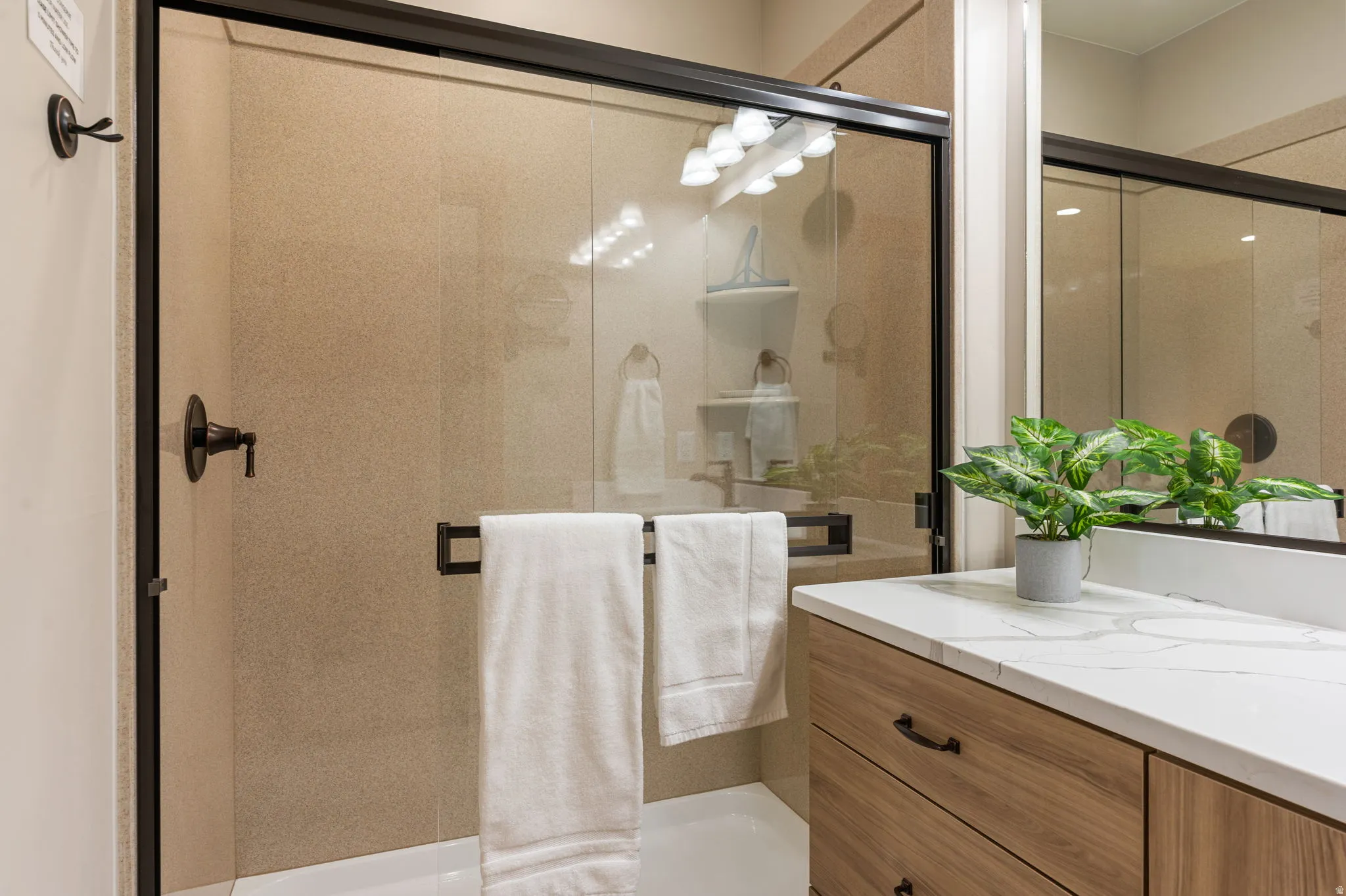 Frameless glass shower enclosure with bronze-finish hardware, light-colored shower surround, wood-finish vanity cabinetry, and a white countertop with gray veining