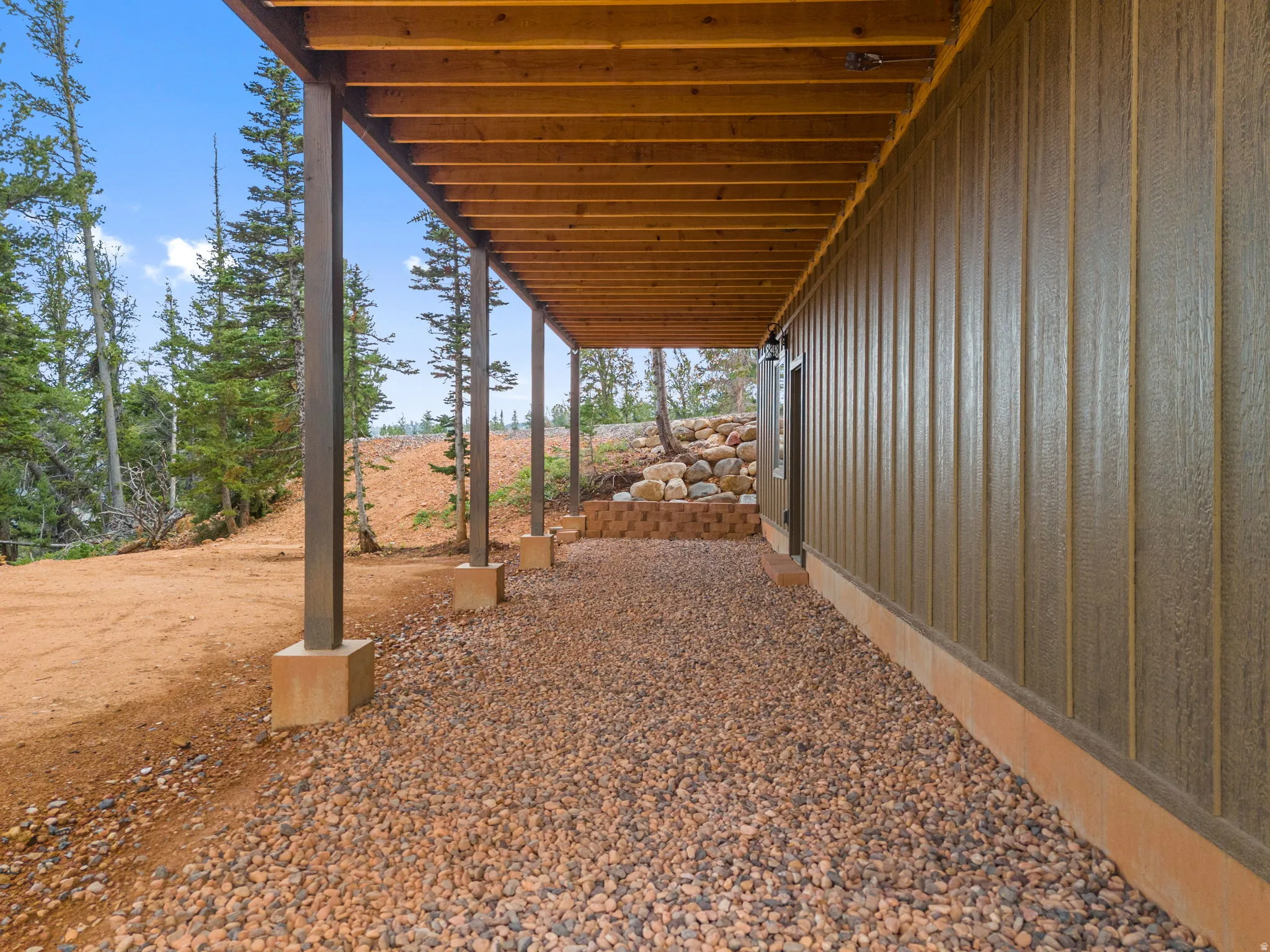 Covered outdoor area featuring exposed wood ceiling joists, support columns with concrete bases, and a gravel surface