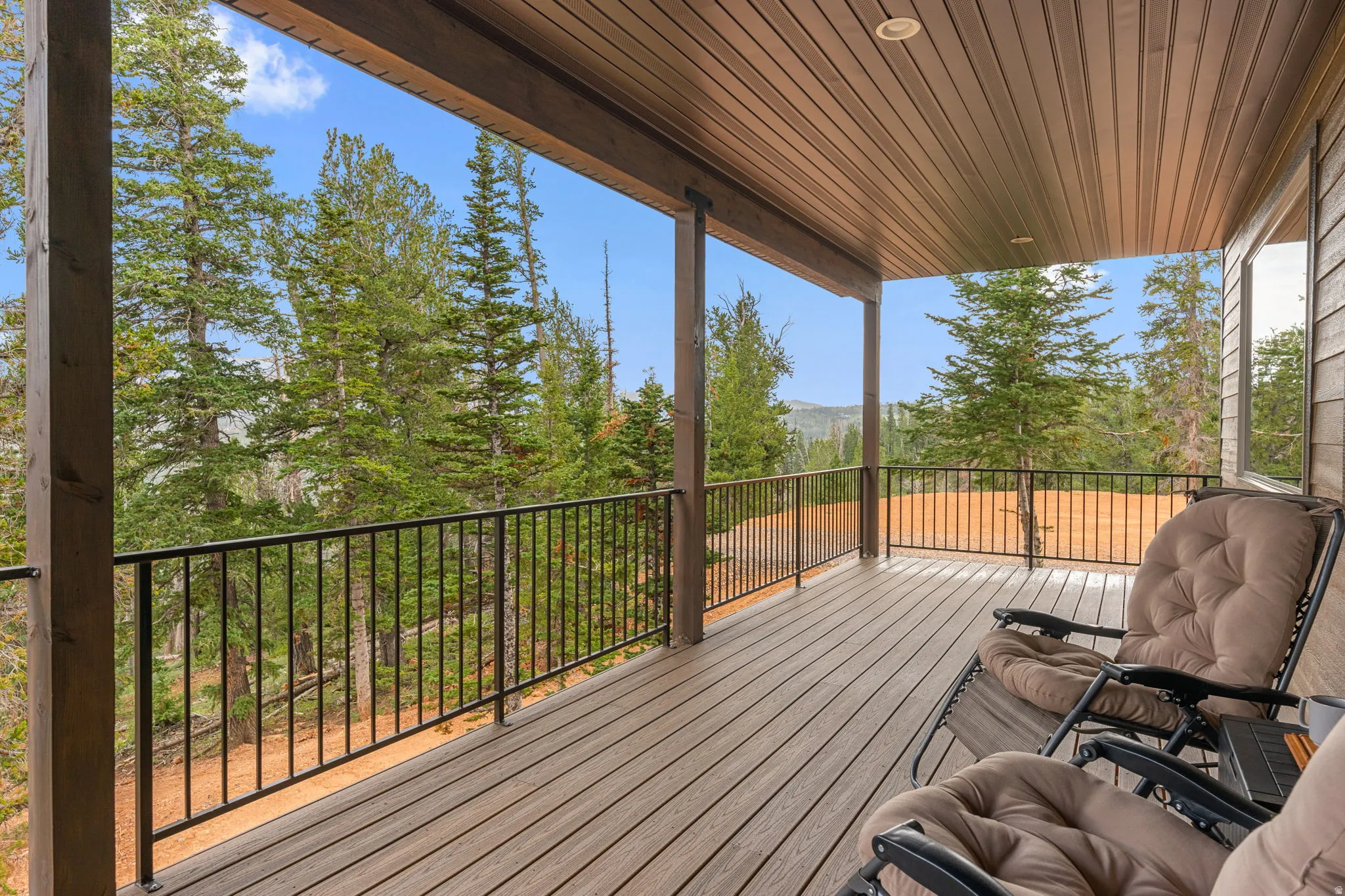 Expansive covered deck with wood-finish decking and black metal railings, set against a backdrop of mature trees and distant hills