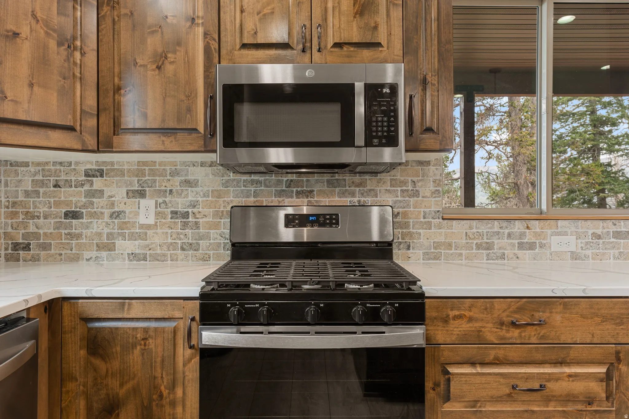 Stainless steel appliance suite, wood cabinetry, stone tile backsplash, white countertops, and a large window