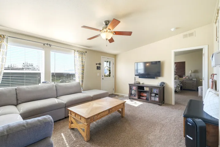 Spacious living area featuring carpet flooring, multiple windows providing natural light, a ceiling fan with integrated lighting, and a white entry door with a decorative glass insert