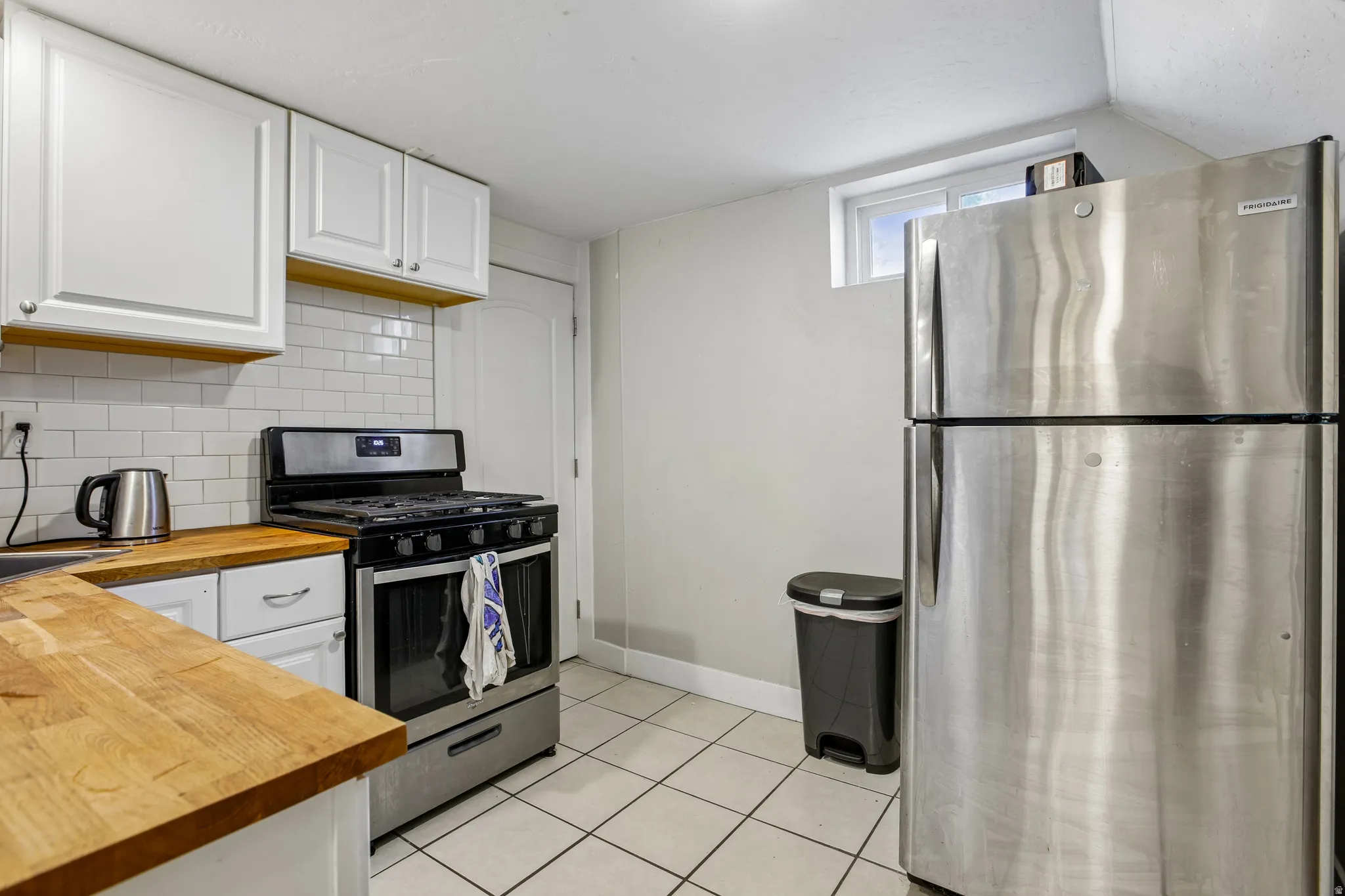 White upper and lower cabinetry, wood-finish countertops, subway tile backsplash, stainless steel gas range, and stainless steel refrigerator