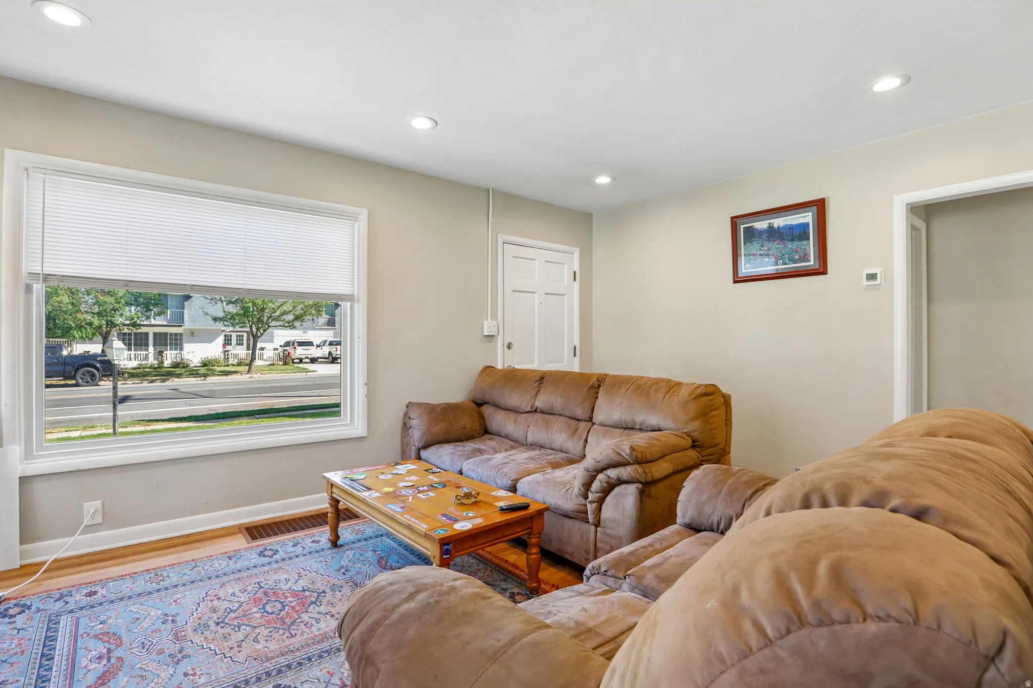 Bright and airy living room featuring large picture windows with cellular shades, recessed ceiling lighting, neutral wall finishes, and hardwood flooring anchored by a patterned area rug